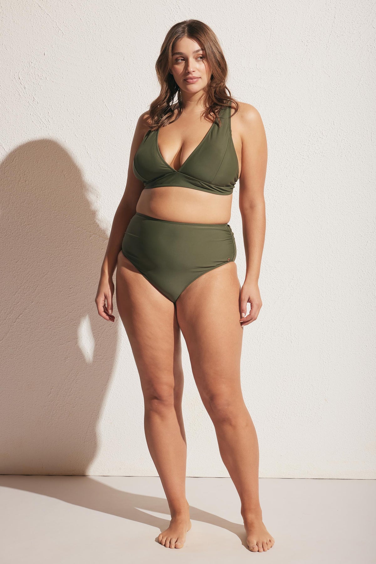 Woman wearing a forest green matte bikini set with a gathered high-waisted bottom, standing barefoot against a plain wall