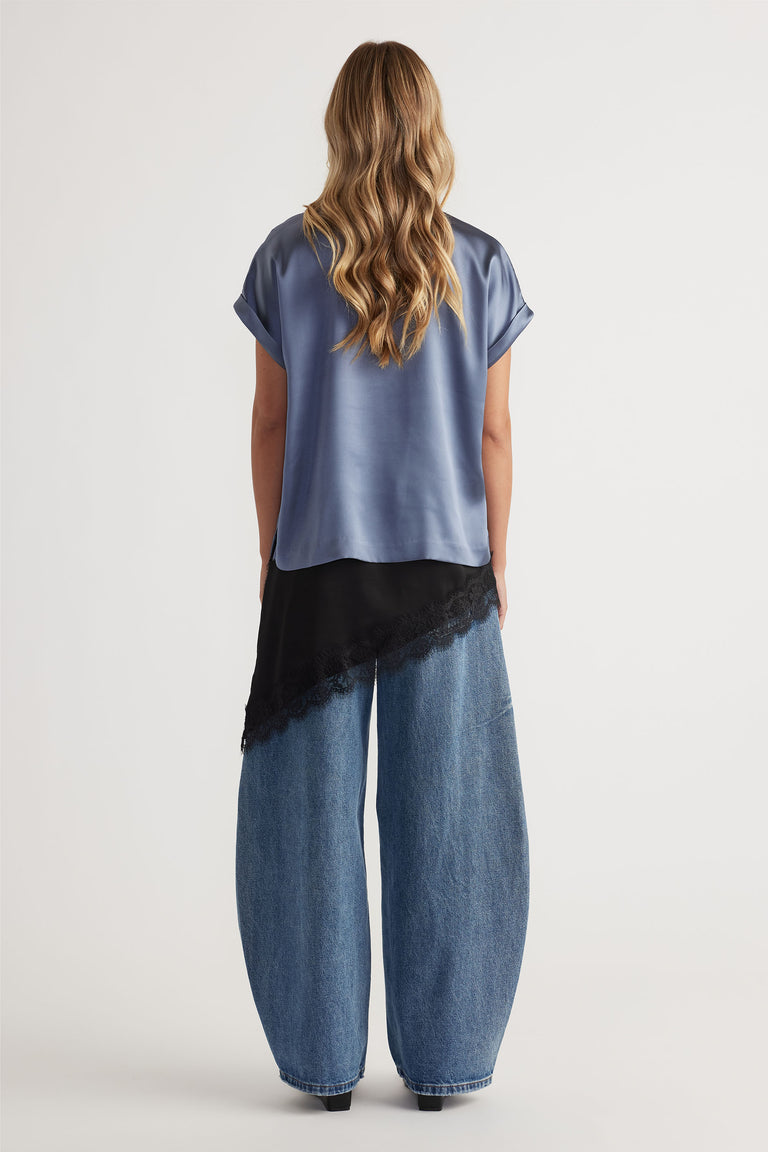 Woman wearing a steel blue satin tee with cuffed sleeves, paired with wide-leg blue jeans and a black lace-trimmed layering top.