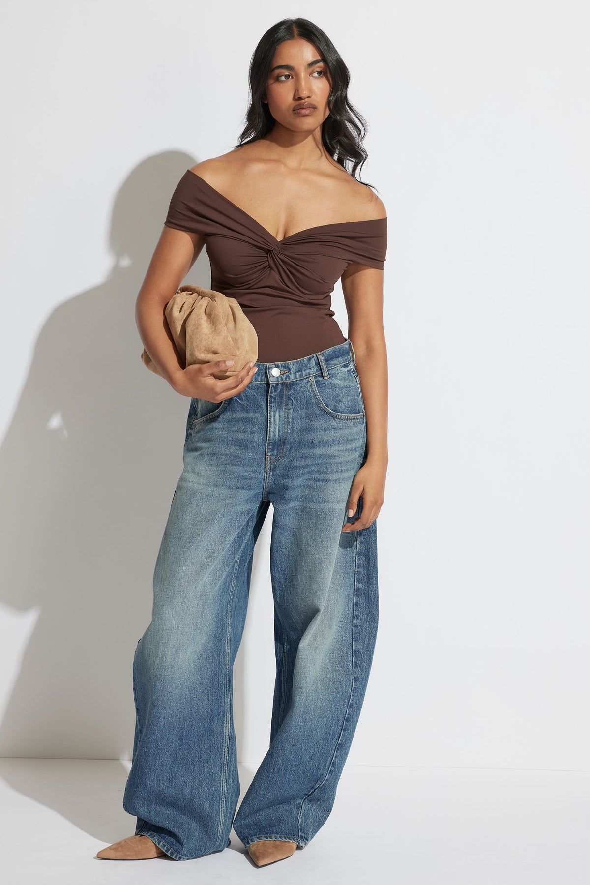Woman wearing Amy Off Shoulder Twist Top in brown with wide-leg jeans, holding a tan clutch in portrait gallery shot.