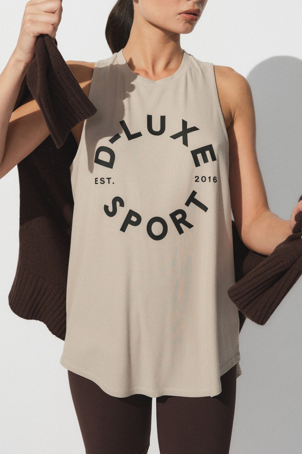 Woman wearing a beige sleeveless tank with bold black circular text paired with dark leggings, styled with a brown knit wrap
