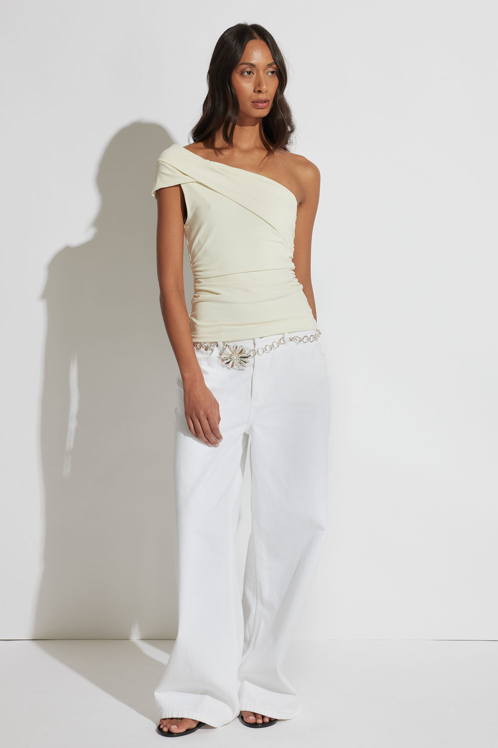 Woman wearing a cream one-shoulder twist top paired with wide-leg white pants and a silver chain belt.