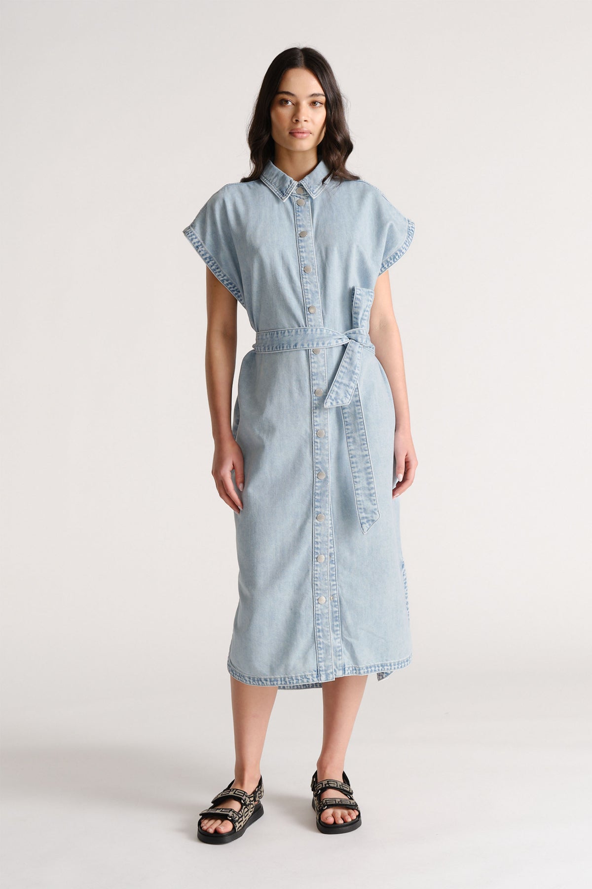 Woman wearing a light blue denim midi dress with a waist tie and cap sleeves, styled with black buckle sandals