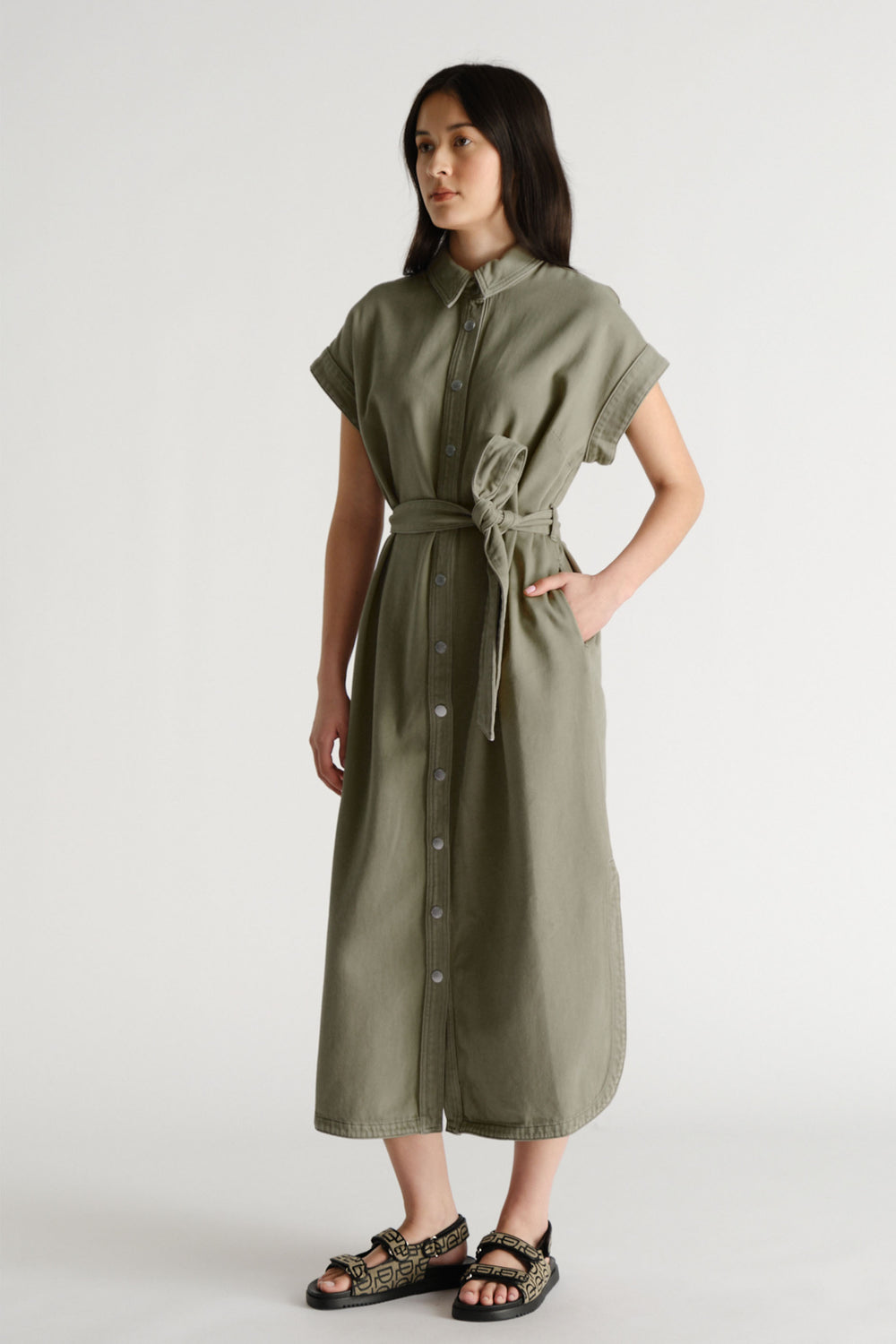 Woman wearing a thyme green button-front maxi dress with rolled sleeves and a matching waist tie belt paired with patterned sandals