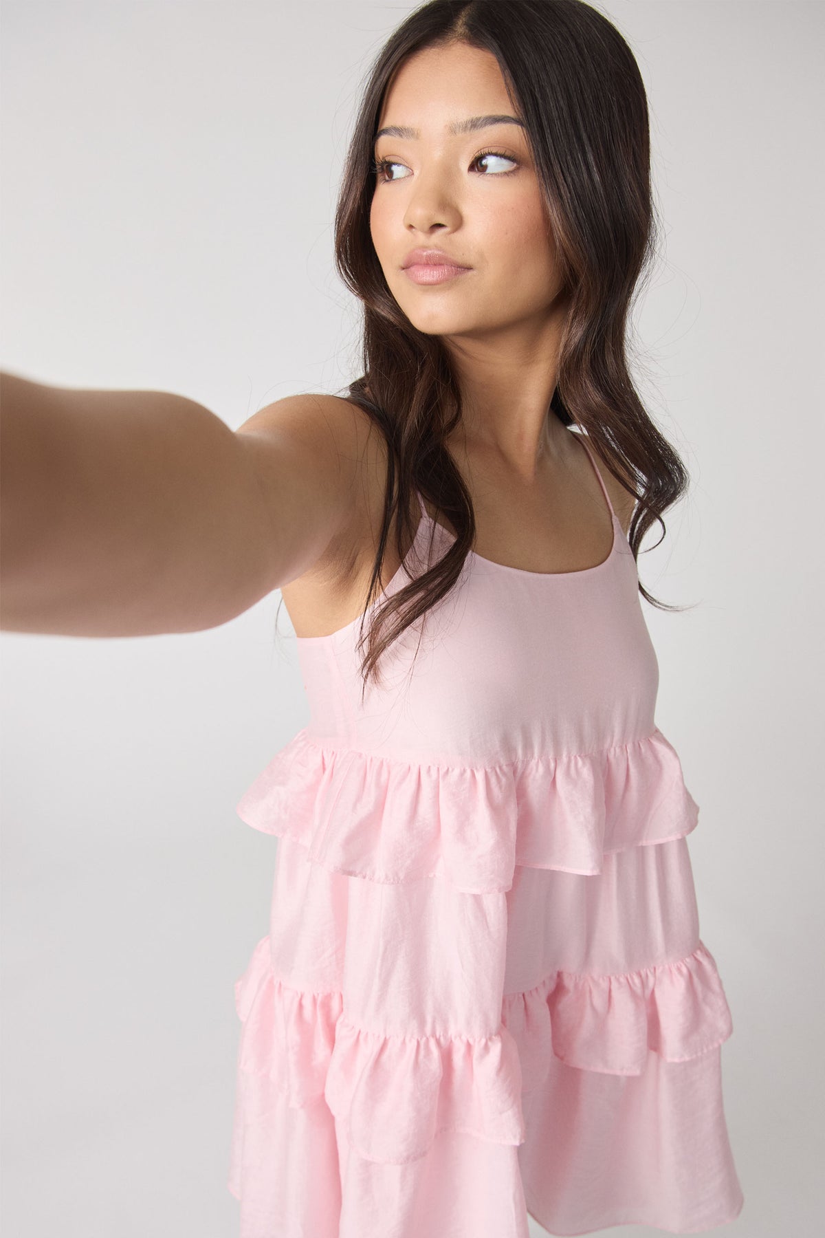 Girl wearing a soft pink ruffle dress with thin straps, taking a selfie against a plain light background.