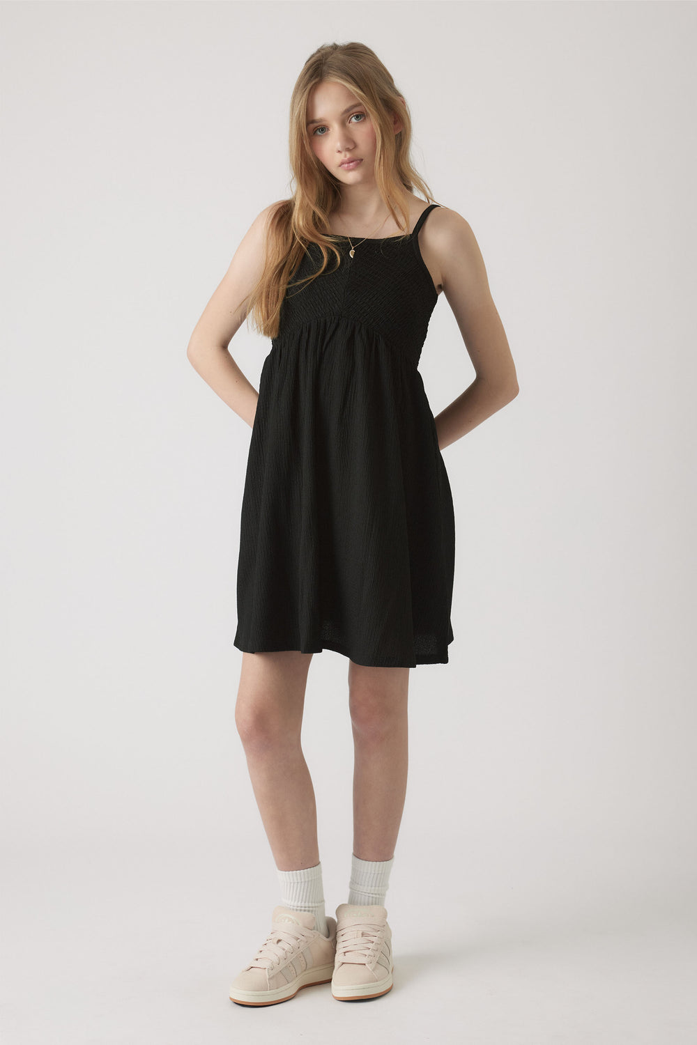 Girl wearing a black shirred dress with thin straps, paired with white socks and cream sneakers in a simple studio setting
