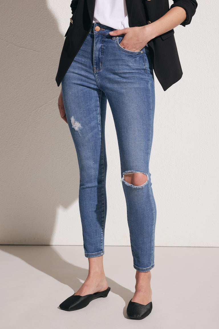 Woman wearing mid-blue distressed skinny jeans with a black blazer and black pointed flats against a neutral background