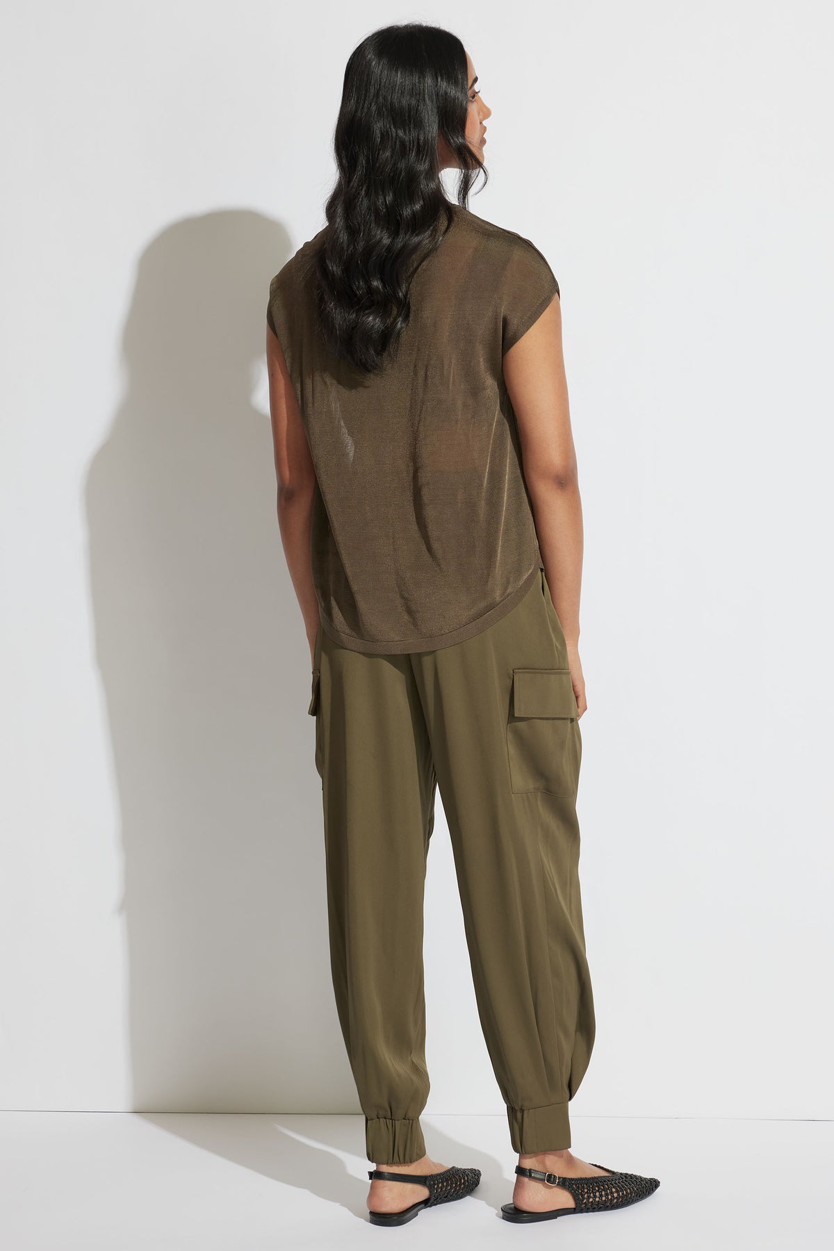 Rear view portrait of model wearing olive Tatum Knit Tee with relaxed fit and sheer fabric, styled with cargo pants and flats