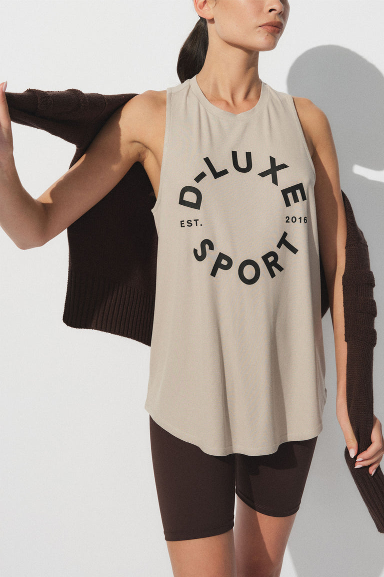 Woman wearing a beige sleeveless tank with bold black lettering paired with dark fitted bike shorts and a sweater draped on shoulders