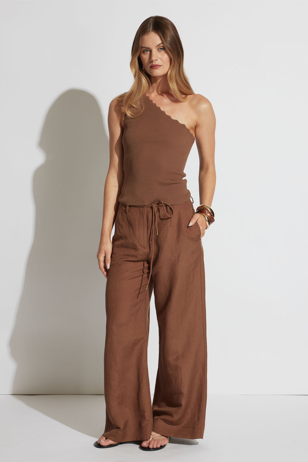 Woman wearing a brown one-shoulder ribbed tank with scalloped edge paired with wide-leg drawstring trousers and bangles
