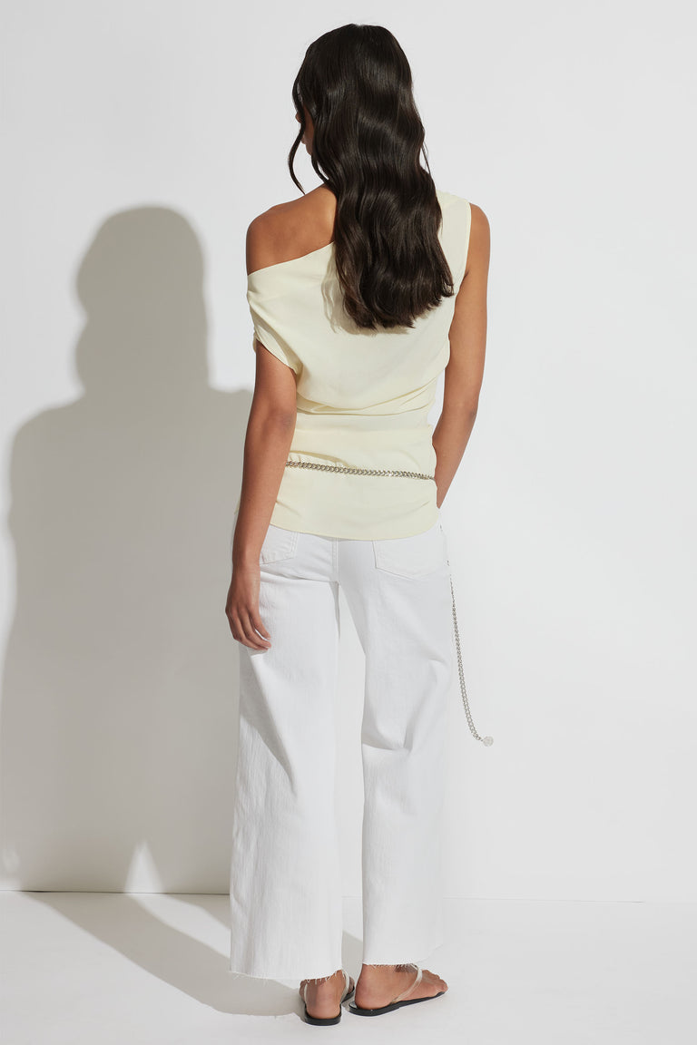 Woman wearing a cream asymmetric shoulder top paired with wide-leg white pants and a silver chain belt accessory