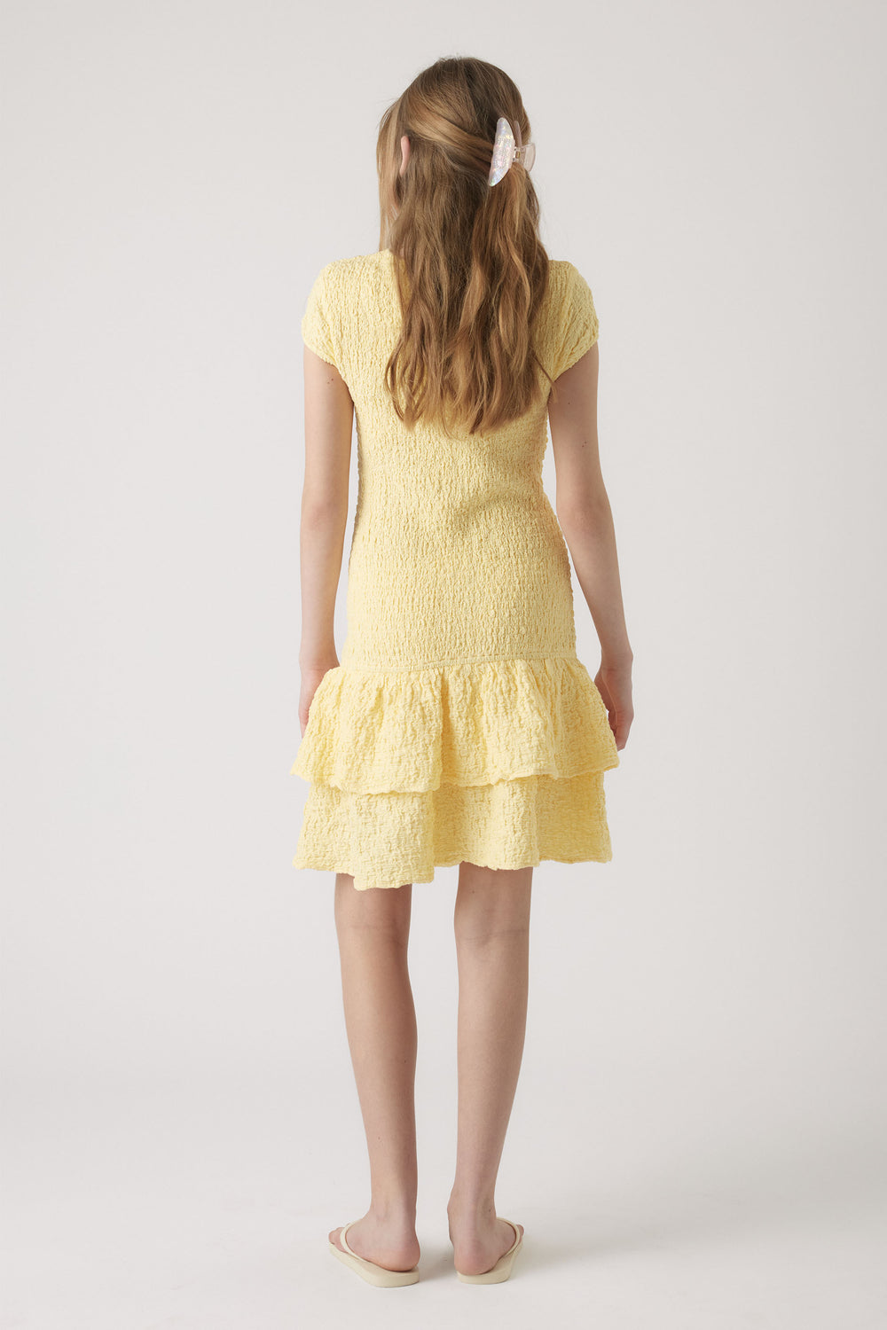 Girl wearing a textured pale yellow shirred dress with short sleeves and a double-layered ruffle hem, paired with beige sandals.