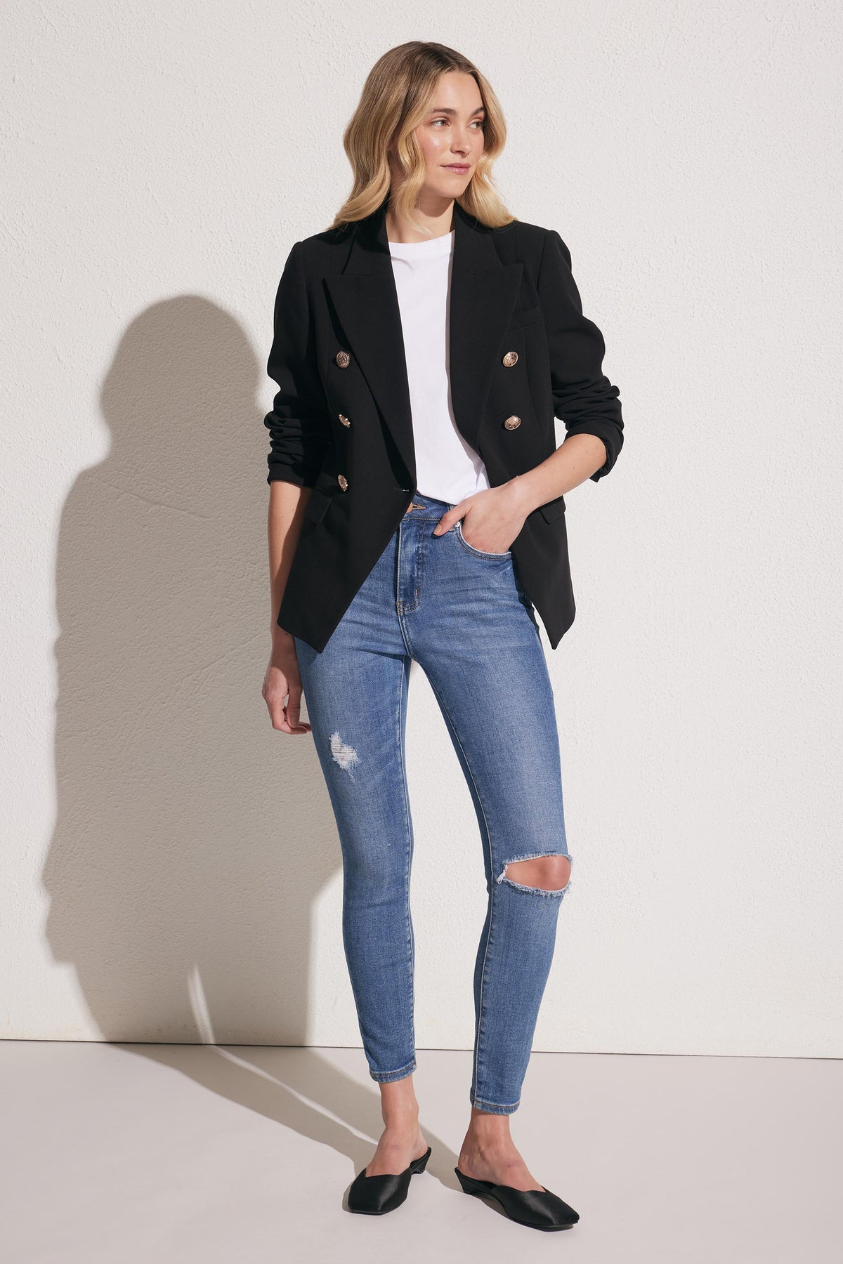 Woman wearing a black double-breasted blazer over a white top with mid-blue distressed skinny jeans and black flats