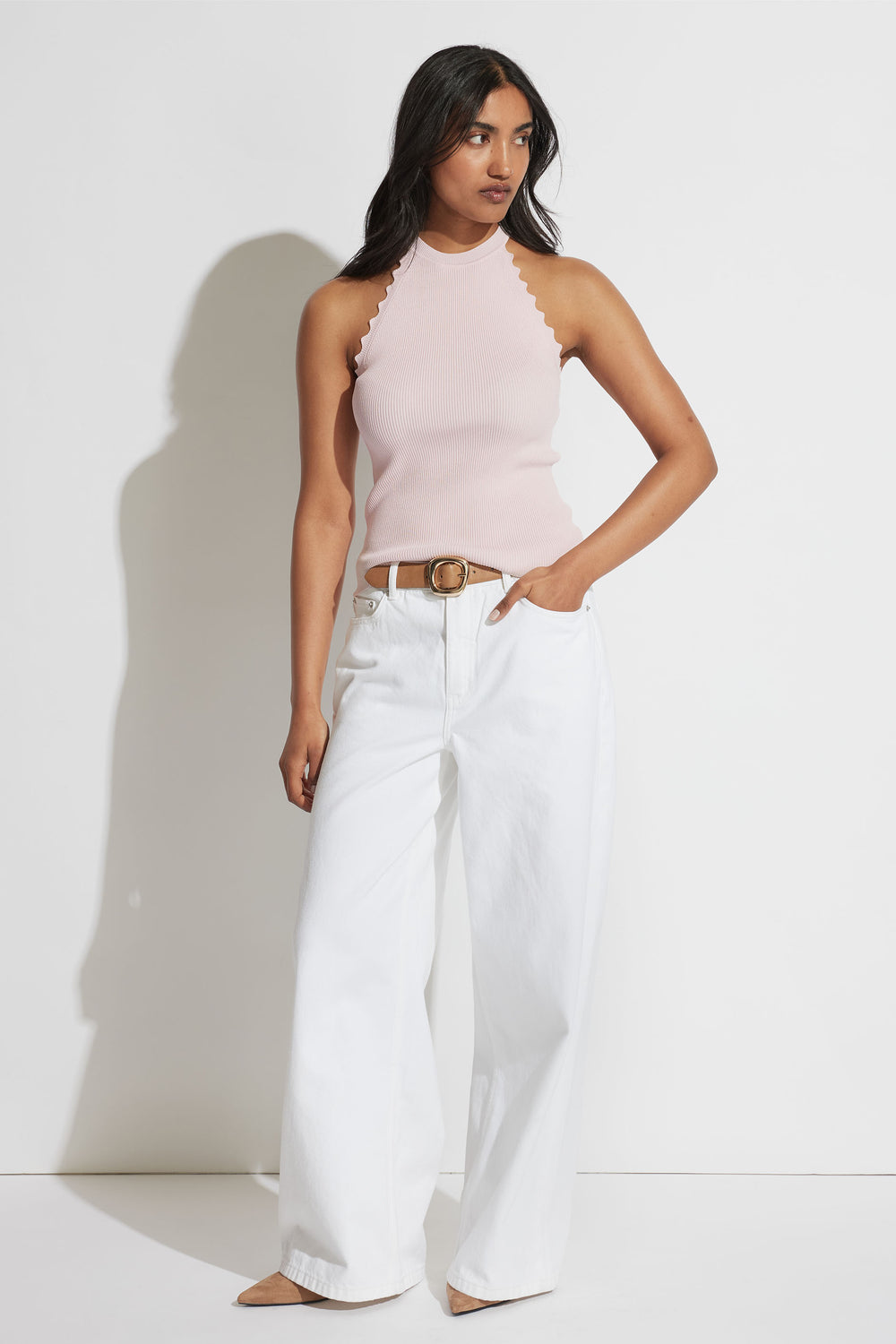 Woman wearing a soft pink scallop-edge halter tank paired with wide-leg white jeans and a tan belt, standing against white wall.