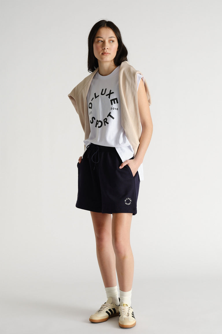 Woman wearing a white graphic tank with a beige sweater draped over shoulders, paired with black shorts and sneakers