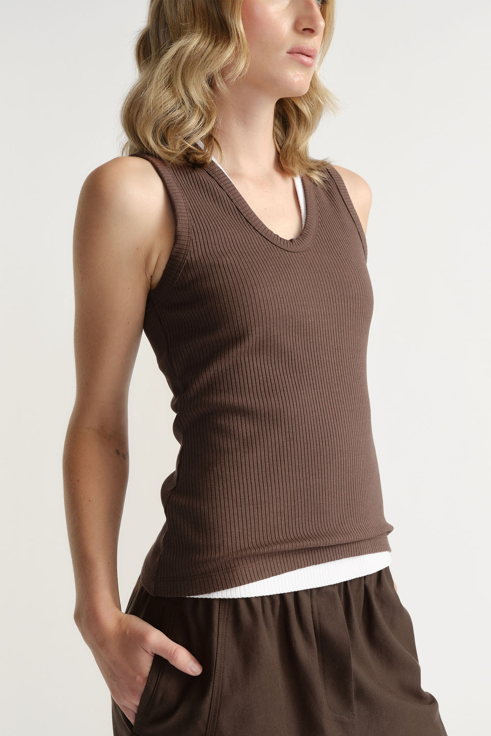 Woman wearing a fitted ribbed scoop neck tank in warm walnut brown layered over a white top with relaxed brown pants