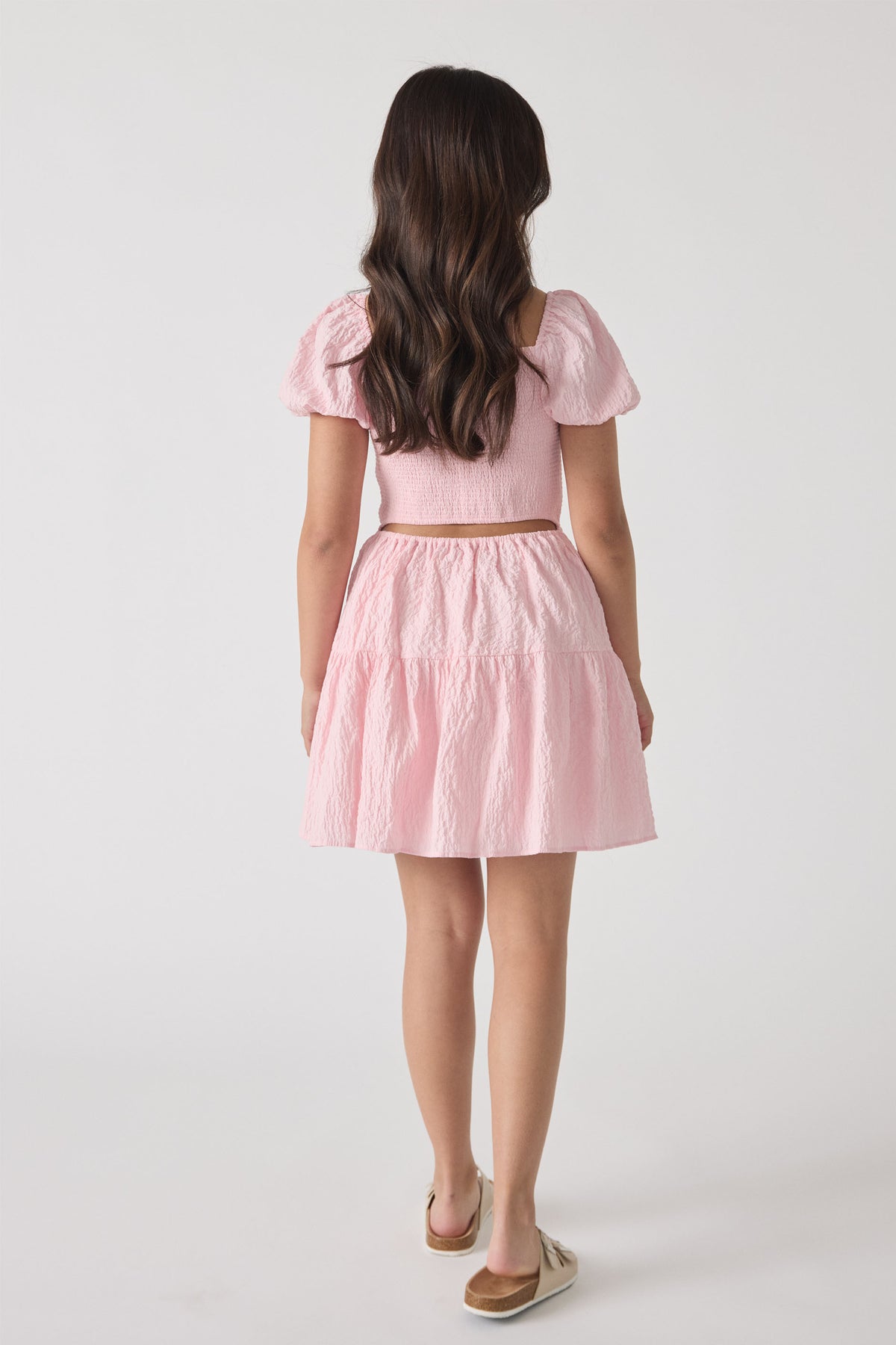 Girl wearing a soft pink tiered mini dress with puff sleeves and textured fabric, shown from behind.