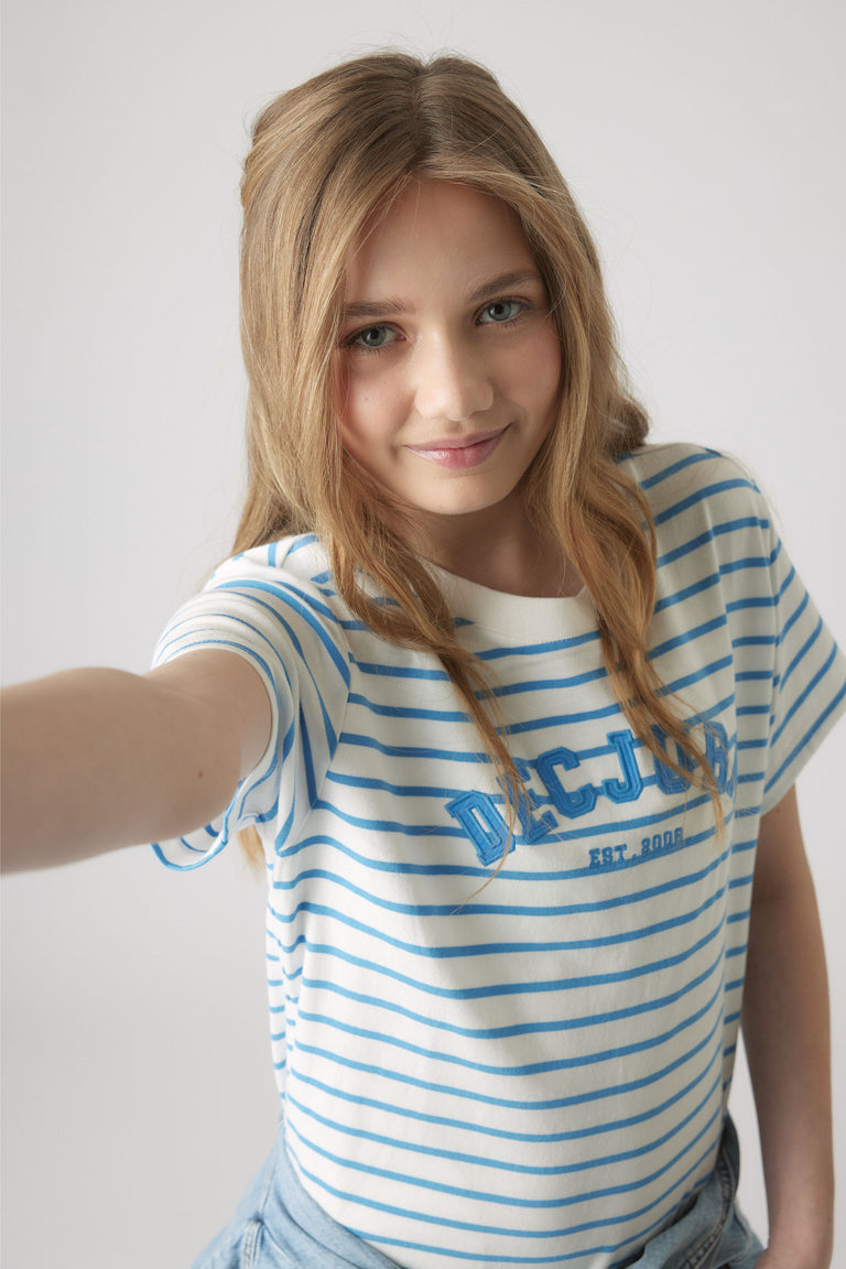 Girl wearing a white and blue striped rugby tee with logo print, styled casually with light denim bottoms against a plain background.