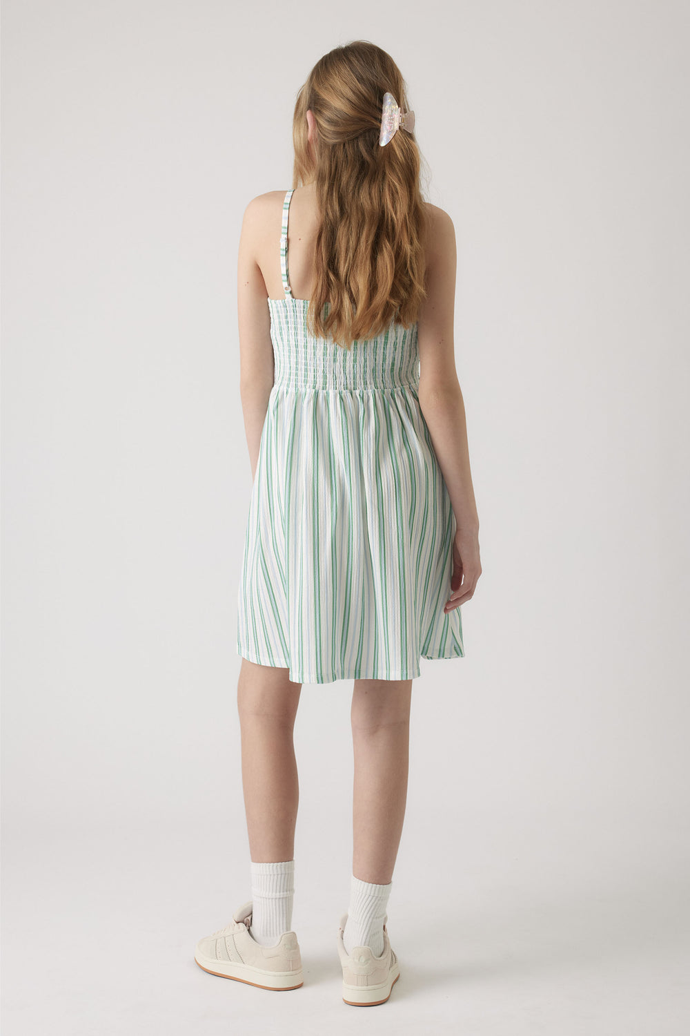 Girl wearing a sleeveless shirred dress with green and white vertical stripes, paired with white socks and sneakers.