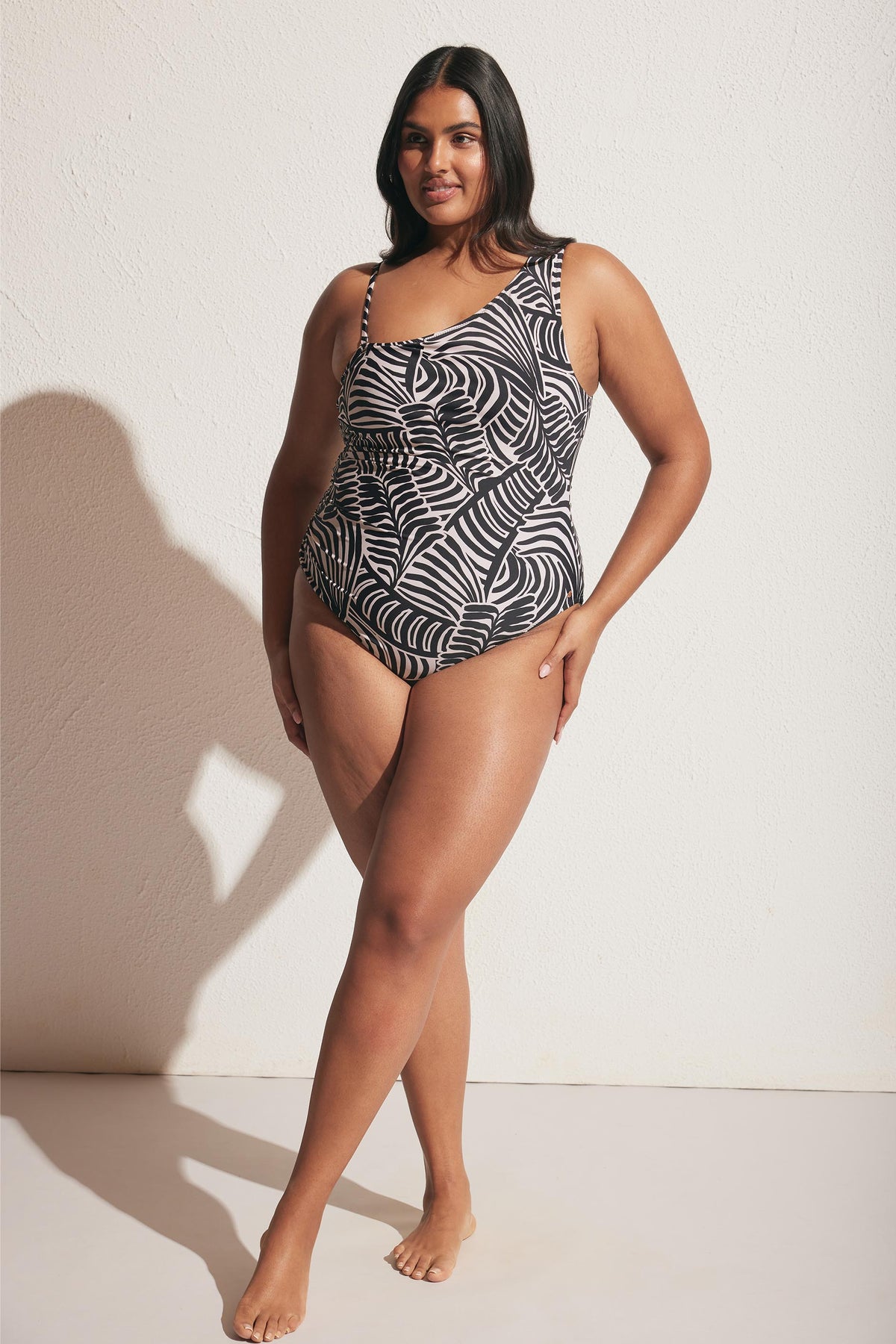 Woman wearing a black and white matte one-shoulder one piece with a bold tropical leaf pattern, standing barefoot.