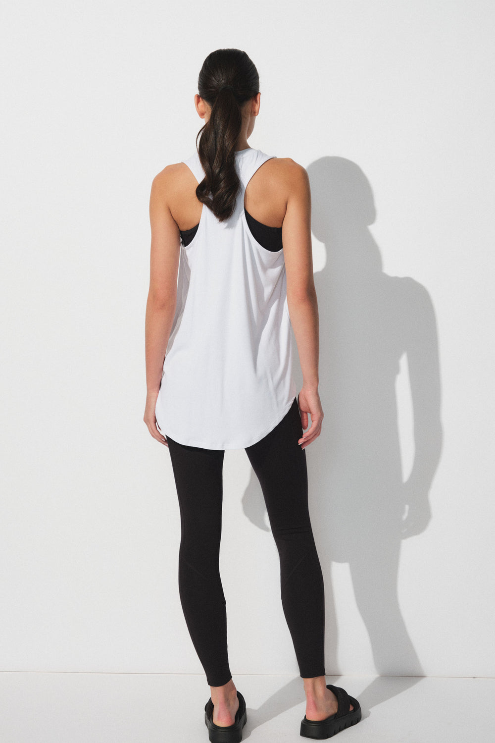 Back view of woman wearing a loose white bamboo tank over a black sports bra paired with black leggings and sandals