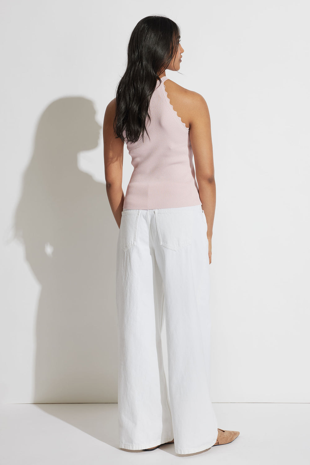 Woman wearing a soft pink scalloped halter tank paired with wide-leg white pants, shown from behind against a white wall.