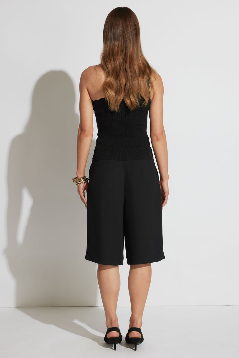 Woman wearing a fitted black one-shoulder tank with loose black knee-length shorts and heeled sandals, shown from behind.
