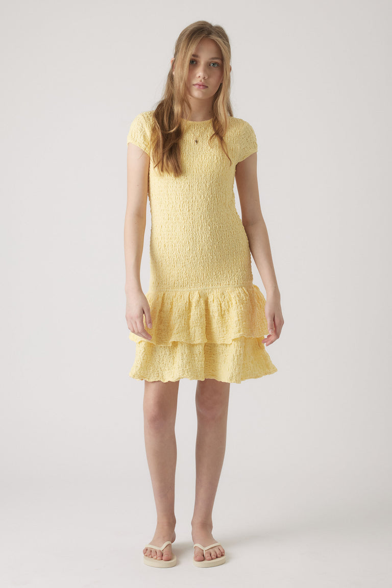 Girl wearing a soft yellow shirred dress with short sleeves and tiered ruffle hem, paired with white sandals
