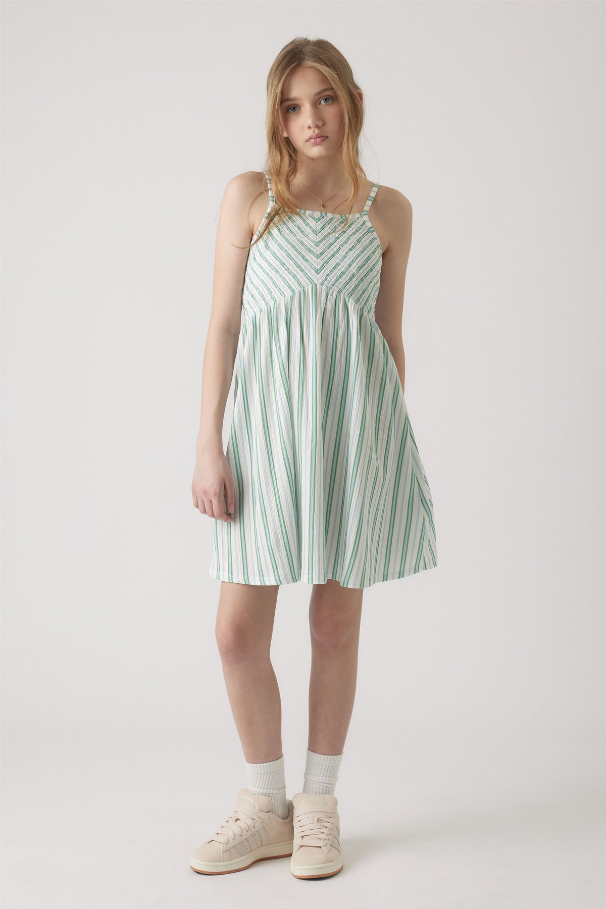 Girl wearing a white and green striped shirred dress with thin straps, paired with white socks and pale sneakers.