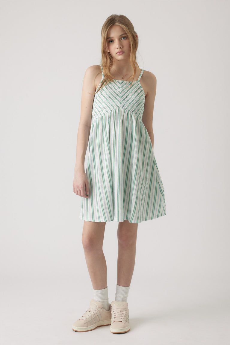 Girl wearing a white and green striped shirred dress with thin straps, paired with white socks and pale sneakers.