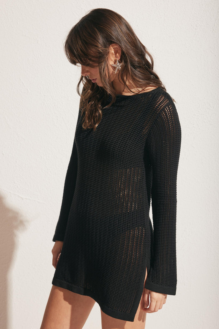 Woman wearing a black crochet tunic with long sleeves and side slits, styled over a black underlayer against a light background