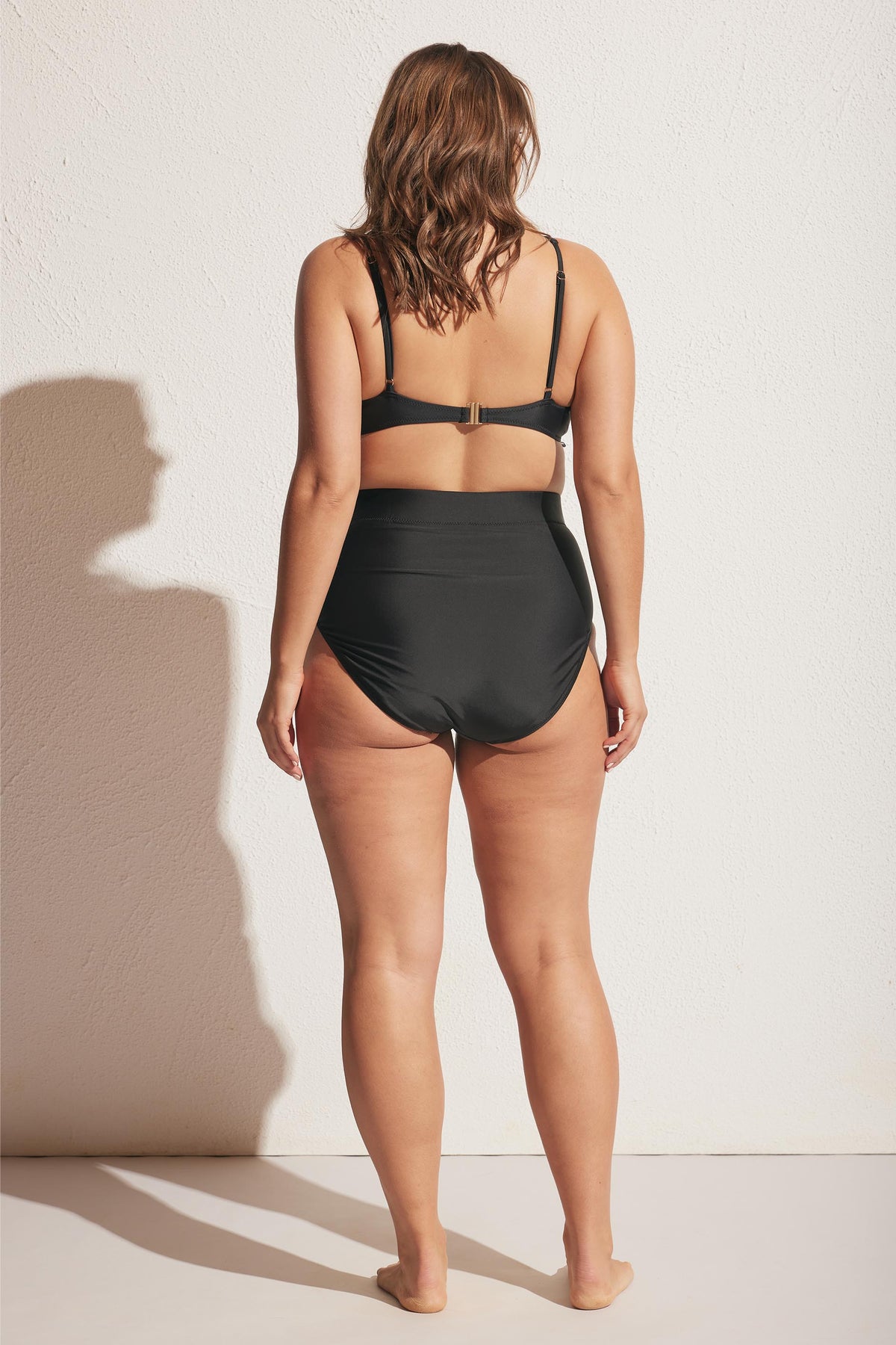Woman wearing a black bikini top with adjustable straps and high-waisted swim bottoms, back view against a plain wall