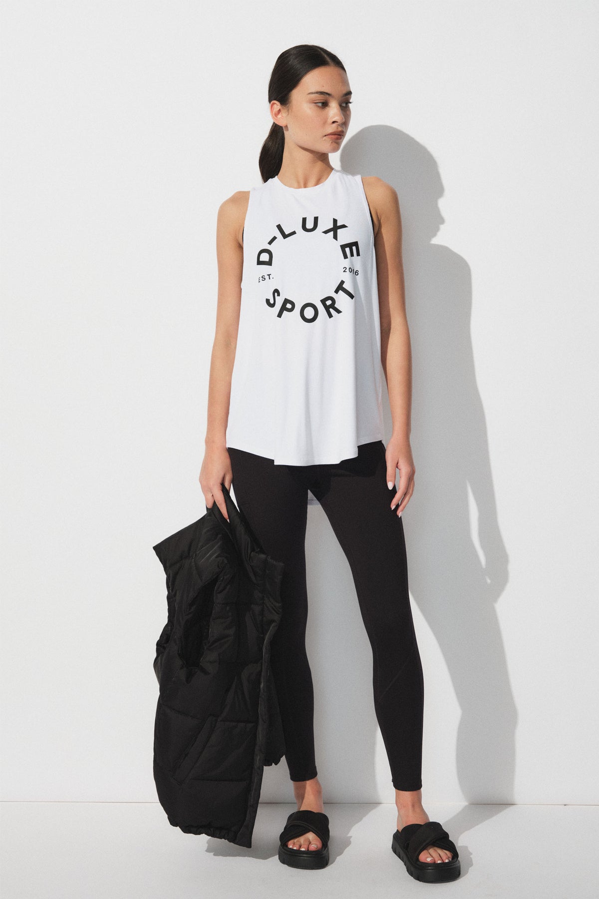 Woman wearing a white sleeveless tank with bold logo print paired with fitted black leggings and black slide sandals