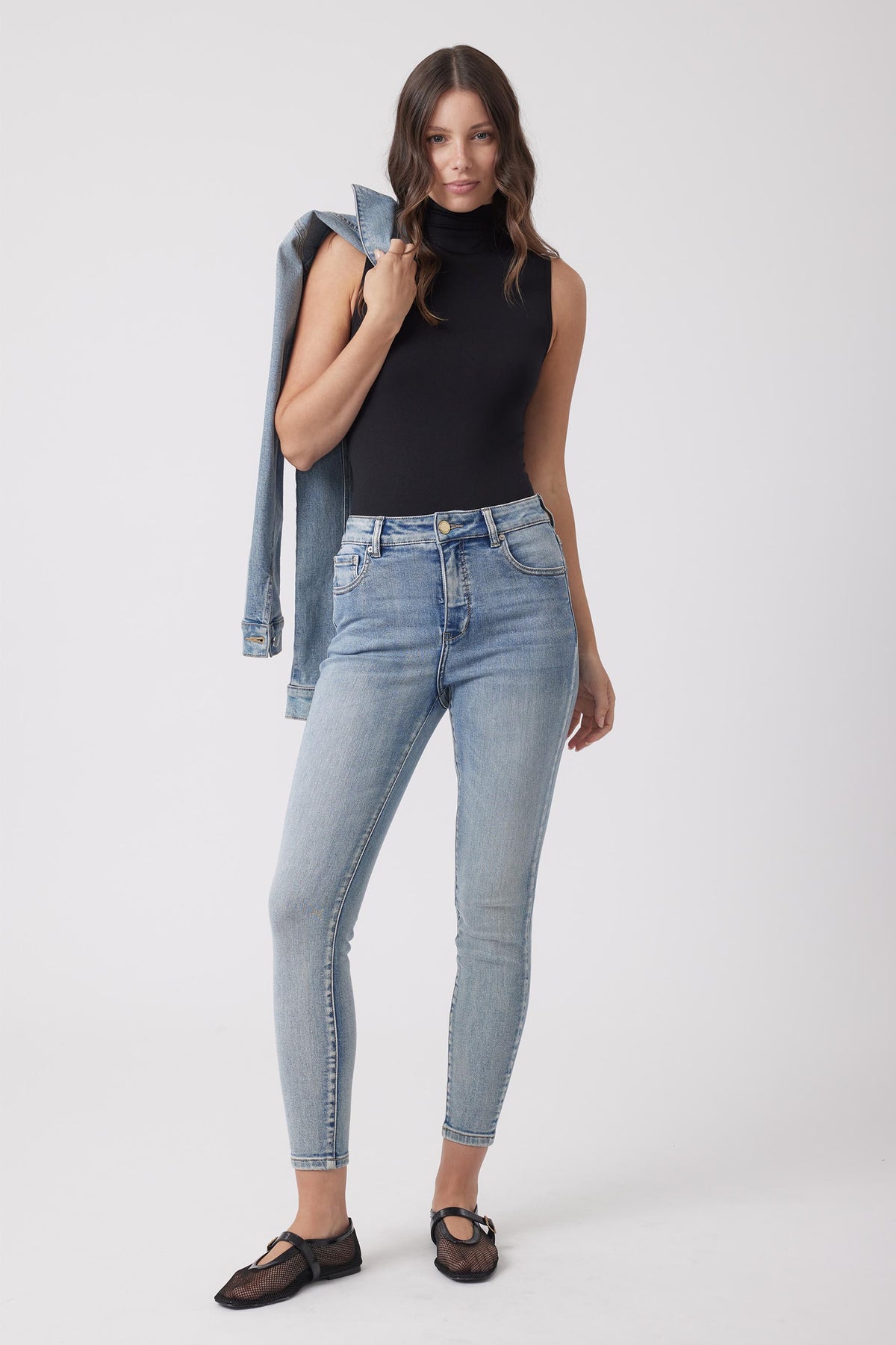 Woman wearing light wash high-rise skinny jeans paired with a fitted sleeveless black turtleneck and holding a denim jacket.