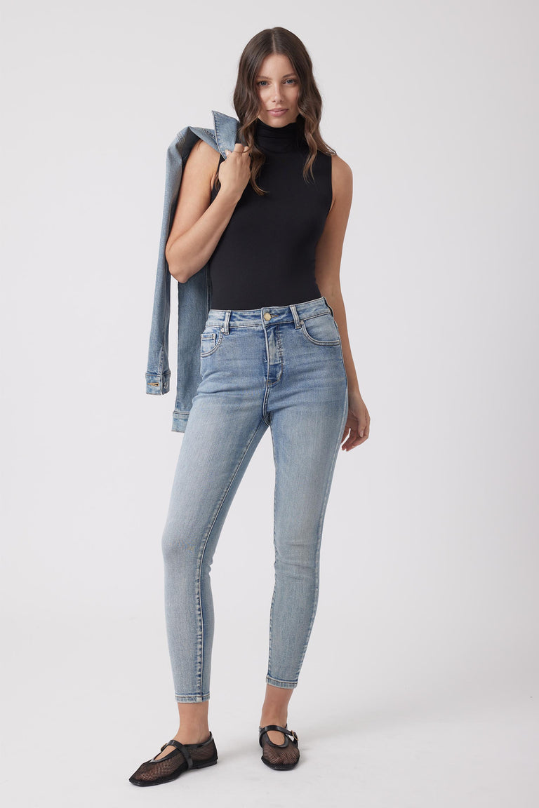 Woman wearing light wash high-rise skinny jeans paired with a fitted sleeveless black turtleneck and holding a denim jacket.