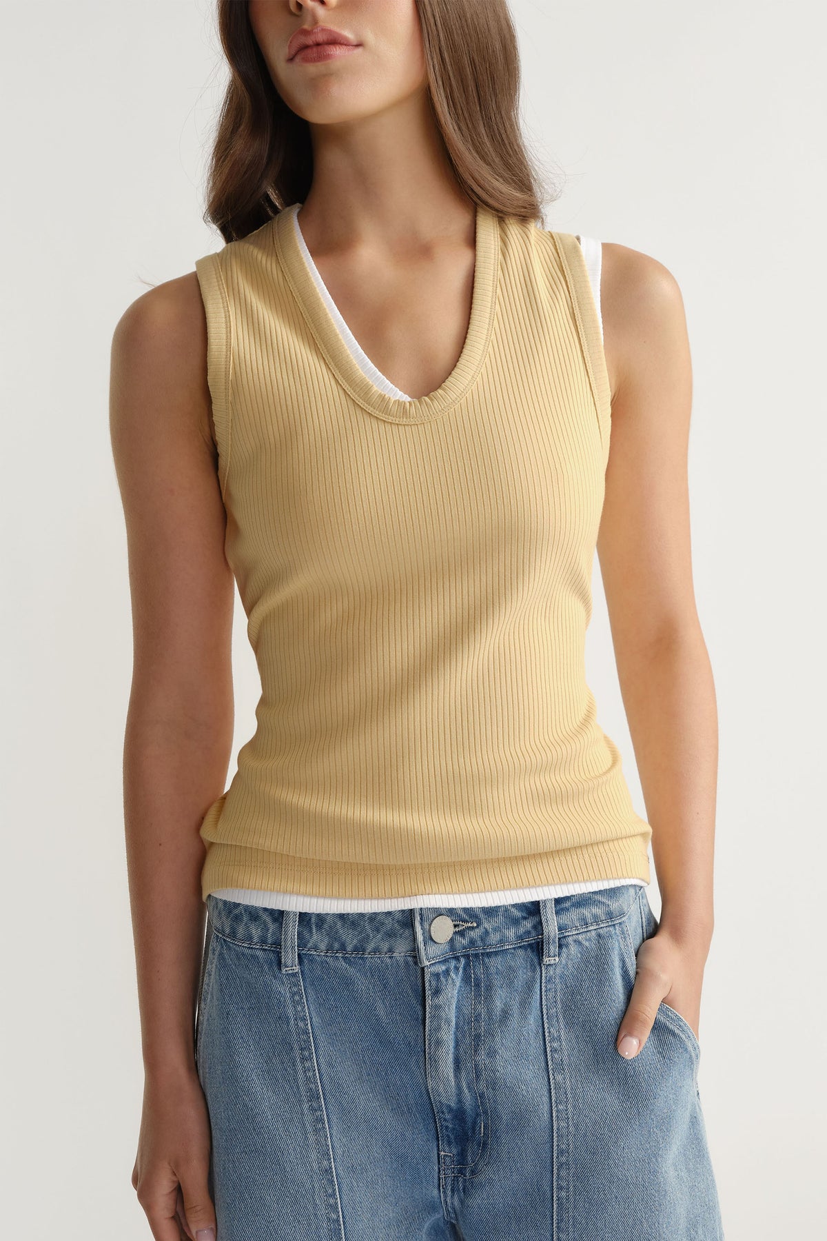 Woman wearing a fitted ribbed light beige scoop neck tank layered over a white top, paired with relaxed blue denim jeans