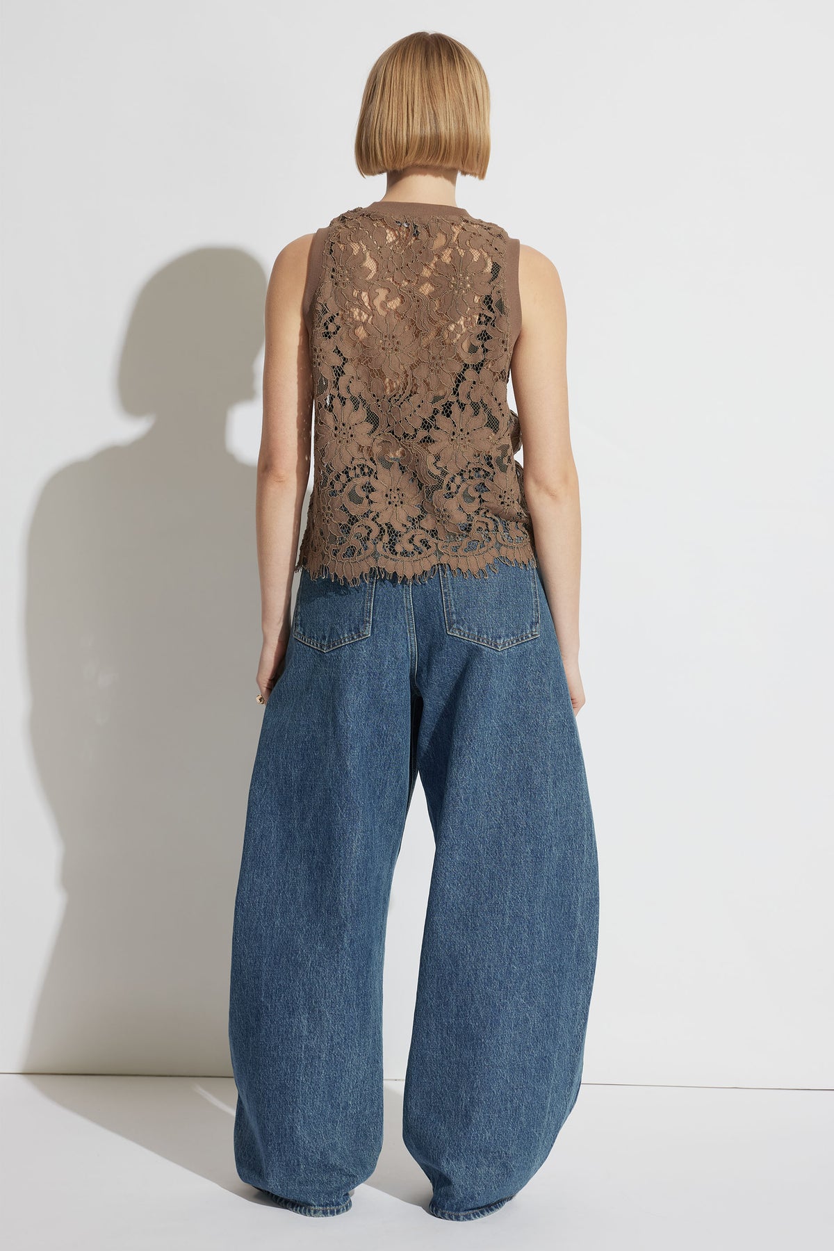 Woman wearing a cappuccino lace tank with floral pattern paired with wide-leg blue denim jeans standing against white background
