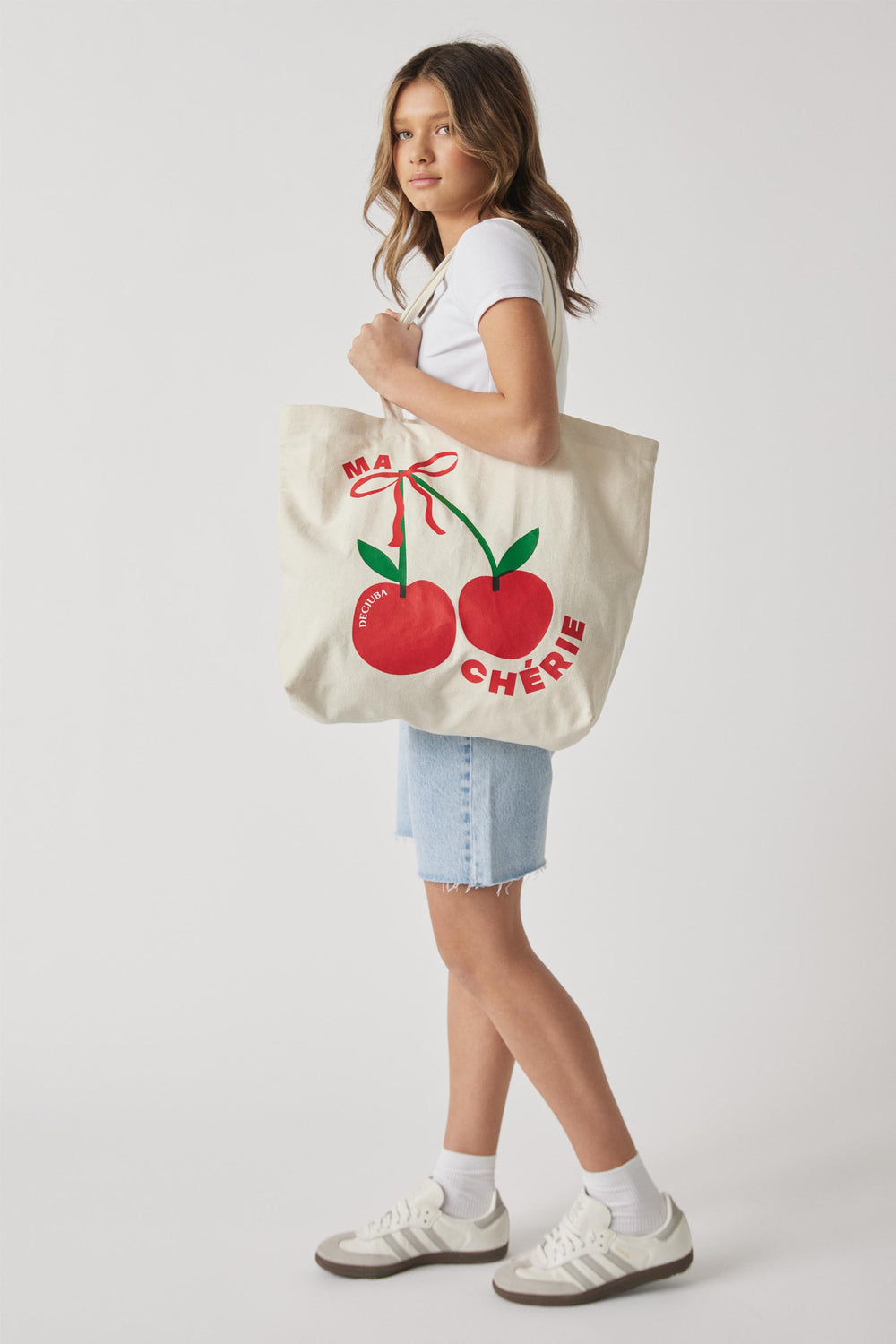 Girl carrying a canvas tote bag with a red cherry graphic, white T-shirt, denim shorts and sneakers