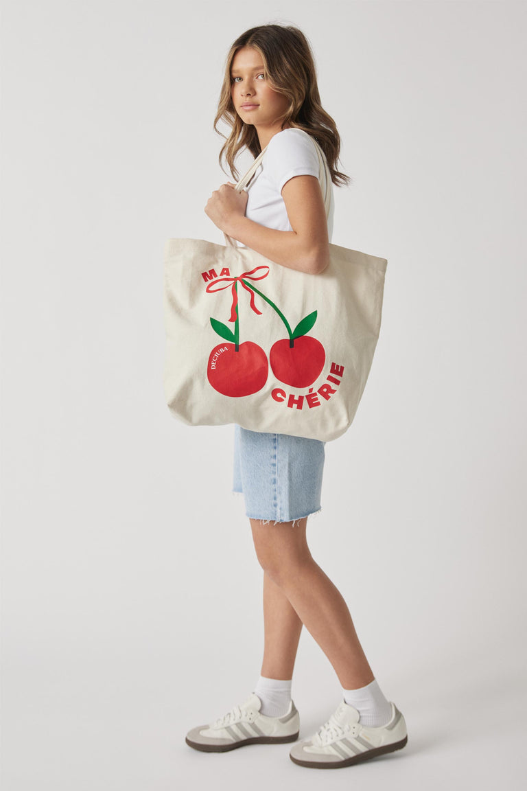 Girl carrying a canvas tote bag with a red cherry graphic, white T-shirt, denim shorts and sneakers