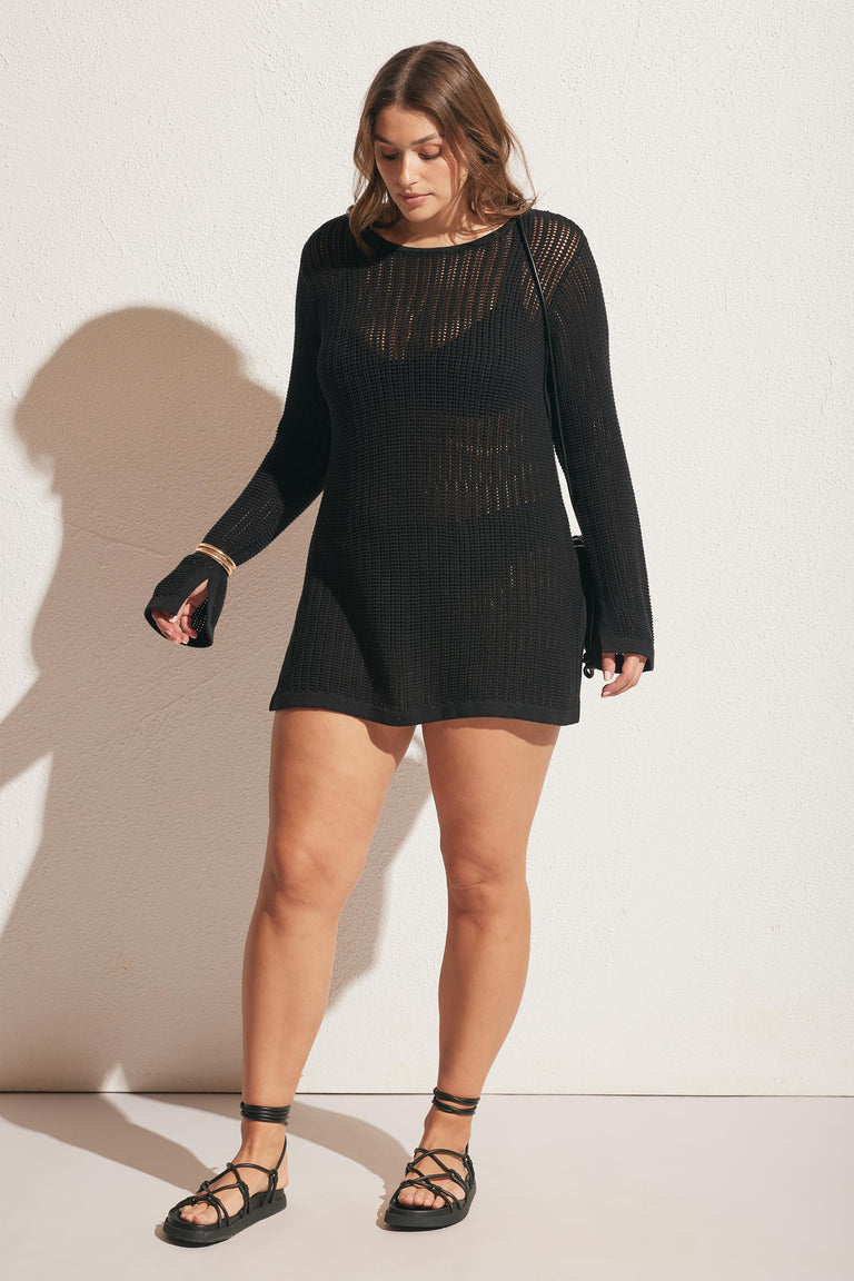 Woman wearing a black crochet tunic dress with long sleeves and lace-up sandals against a plain wall