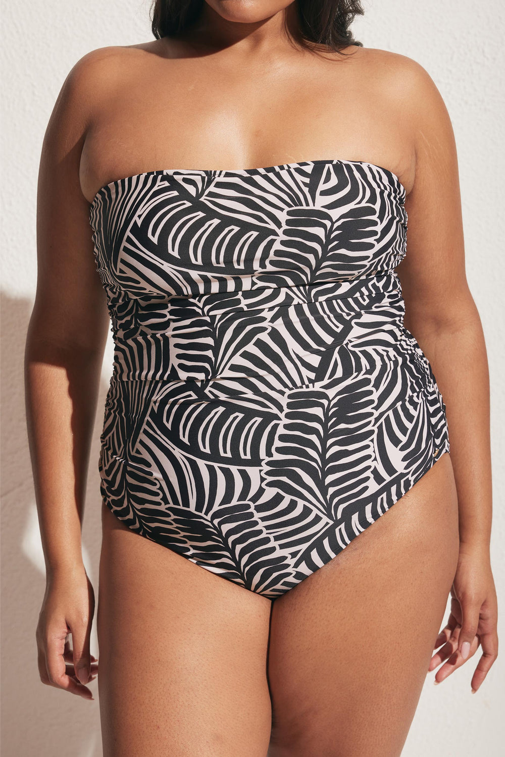 Woman wearing strapless black and white ruched swimsuit with abstract leaf pattern standing against textured wall