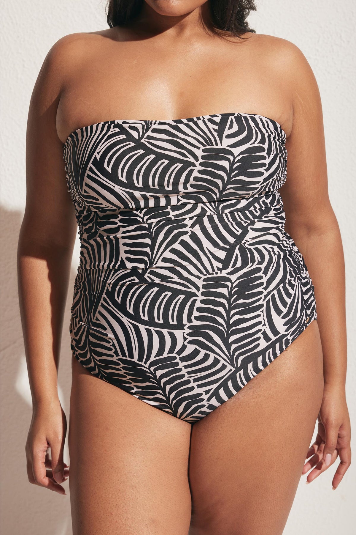 Woman wearing strapless black and white ruched swimsuit with abstract leaf pattern standing against textured wall