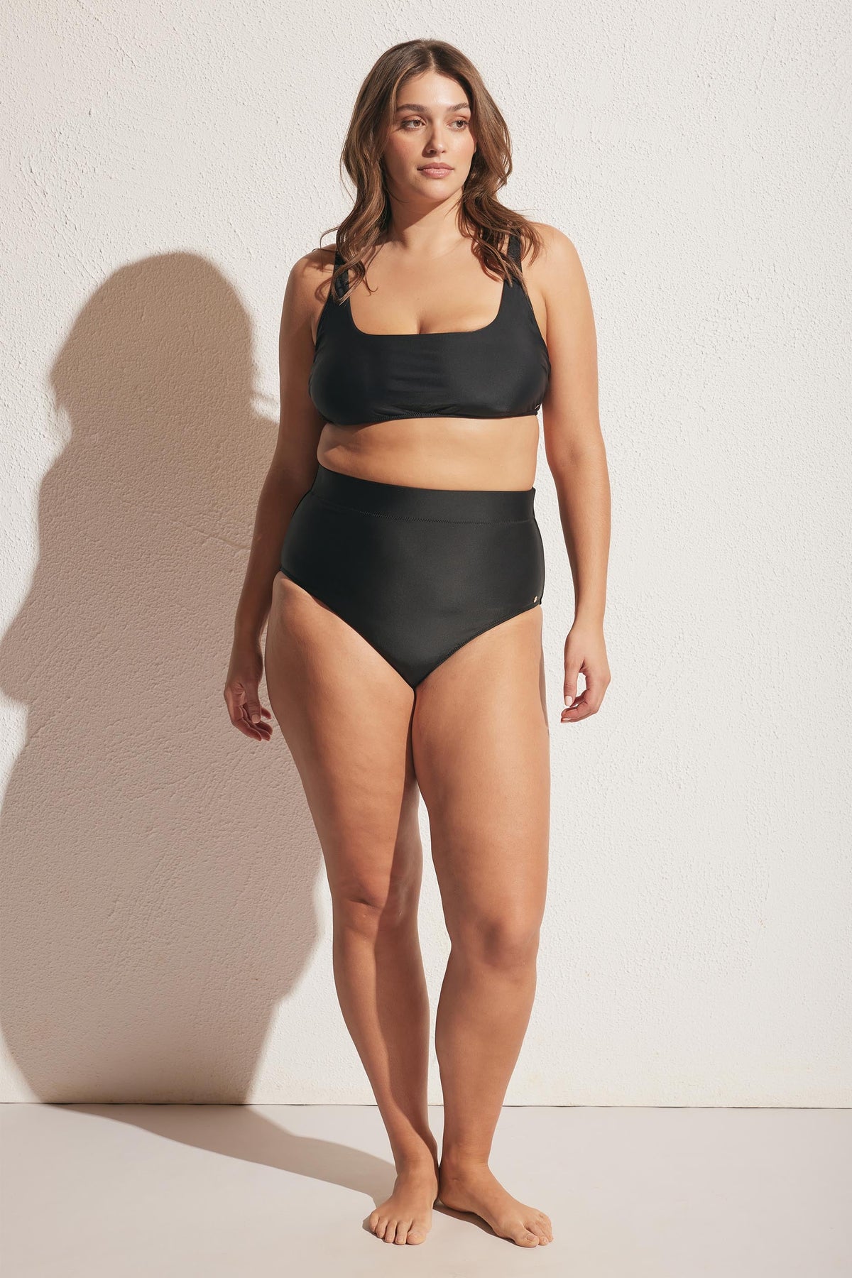 Woman wearing a sleek black high-waisted bikini bottom with a matching crop-style swim top standing against textured light wall