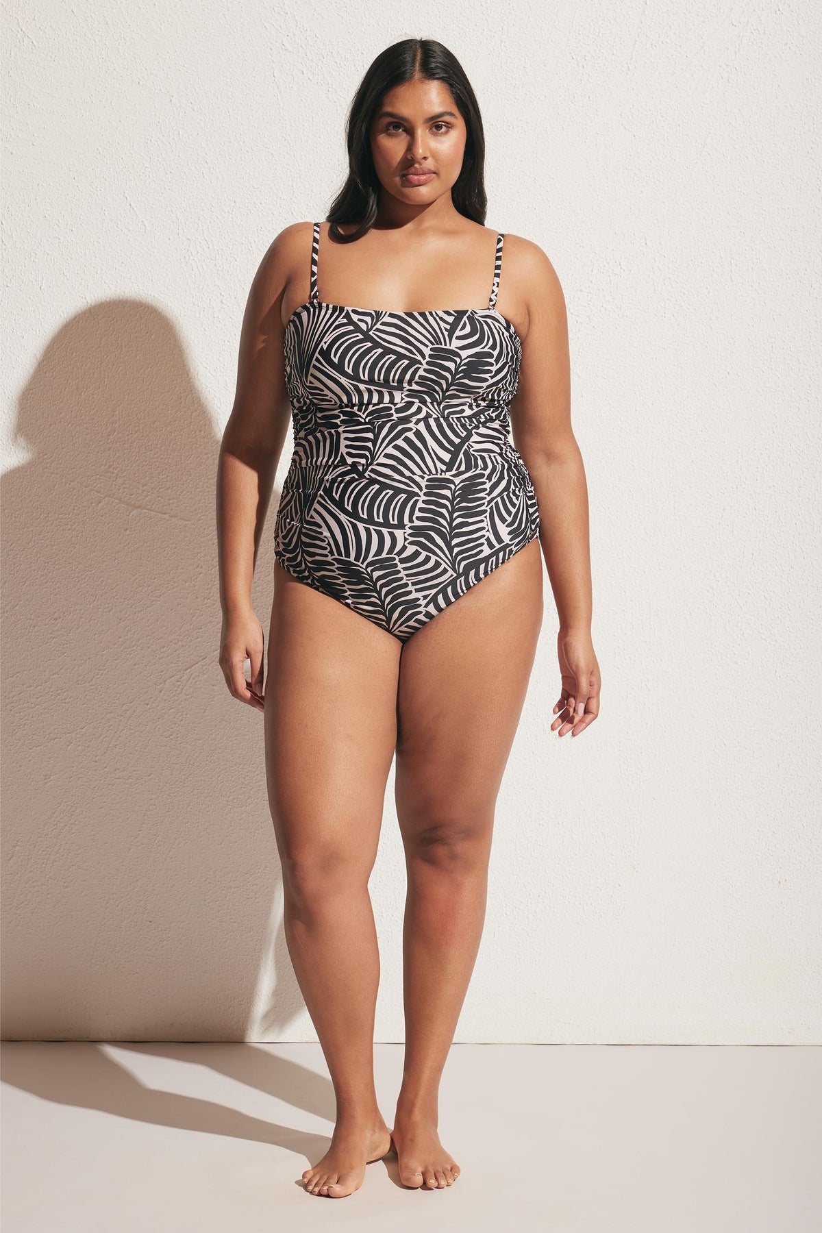Woman wearing a matte one-piece swimsuit with black and white palm leaf print, standing barefoot against a light wall