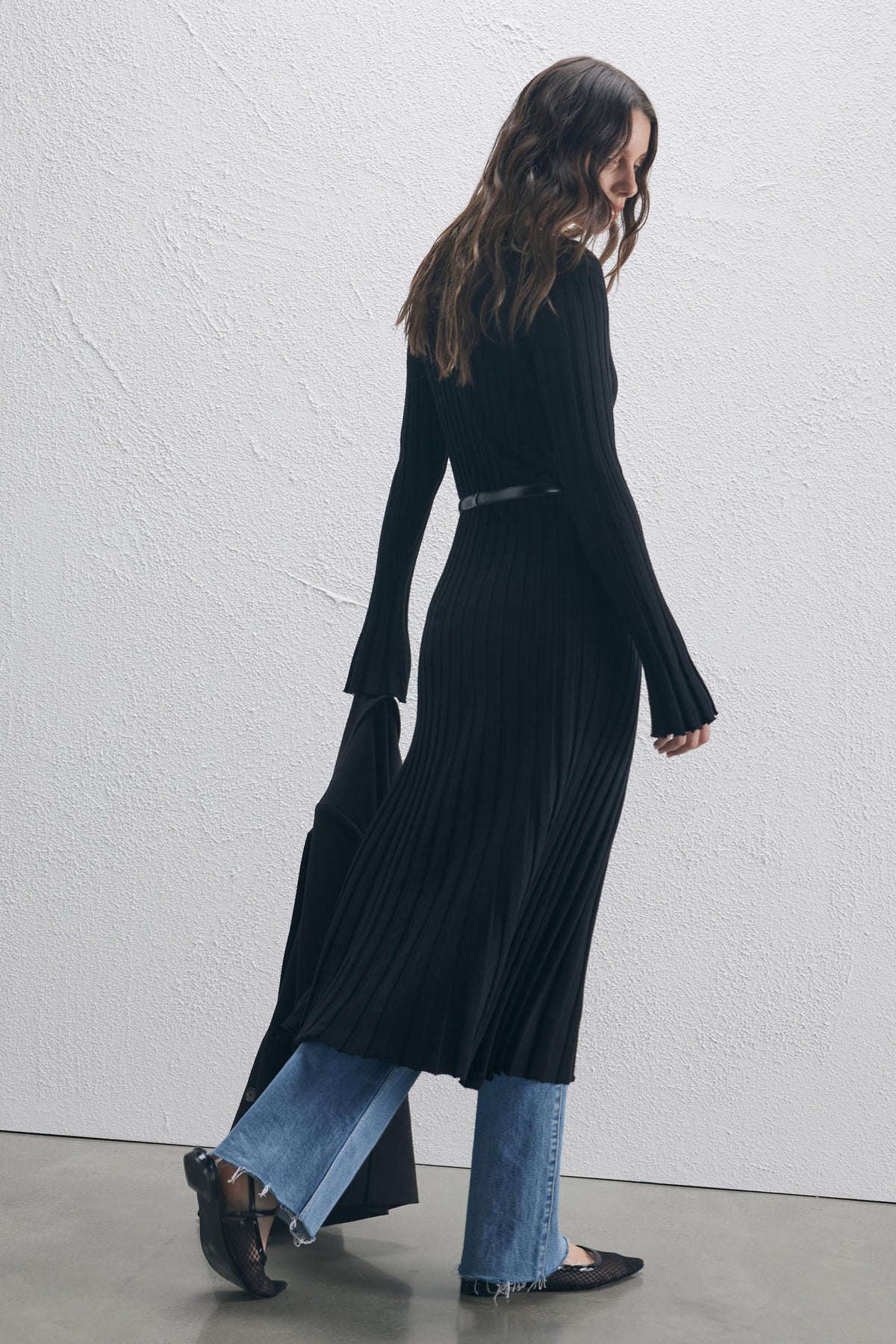 Woman in a black ribbed scoop neck knit dress styled over mid-blue frayed hem jeans and black pointed flats walking indoors
