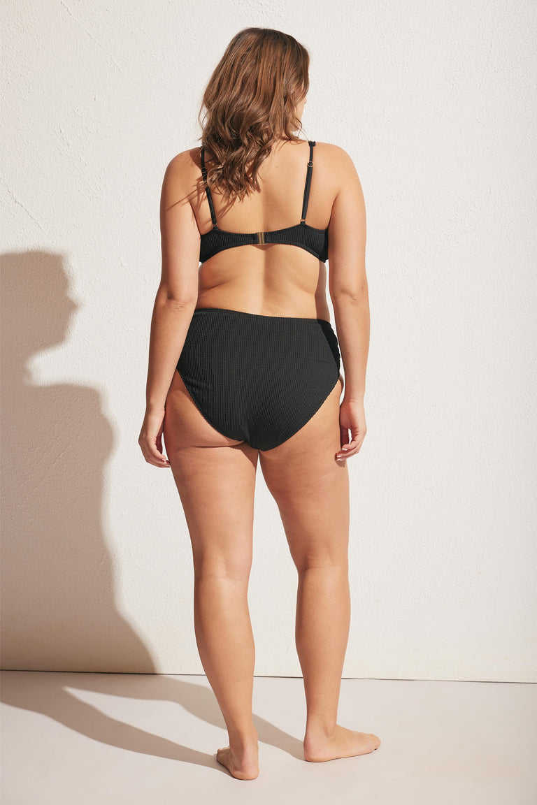 Woman wearing a high-waisted black bikini bottom with a textured crinkle fabric, shown from the back in natural light