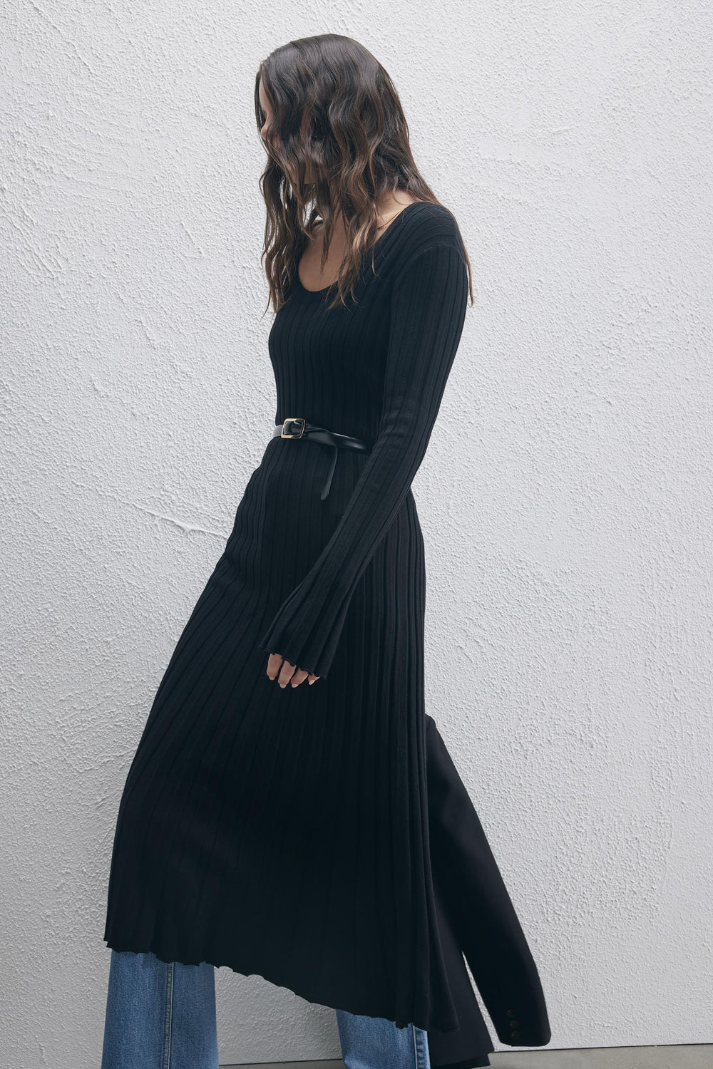 Woman wearing a black ribbed knit scoop neck dress with a belt layered over blue jeans, walking by a textured wall