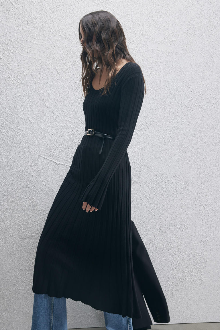 Woman wearing a black ribbed knit scoop neck dress with a belt layered over blue jeans, walking by a textured wall