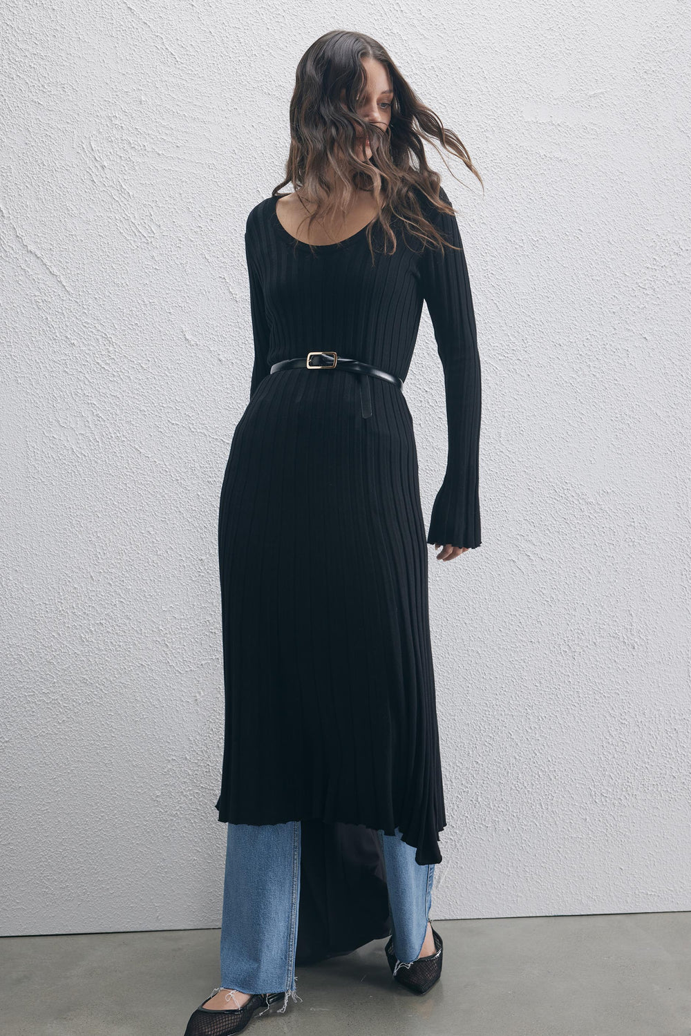 Woman wearing a black ribbed knit scoop neck dress with long sleeves, styled with a belt and layered over blue jeans