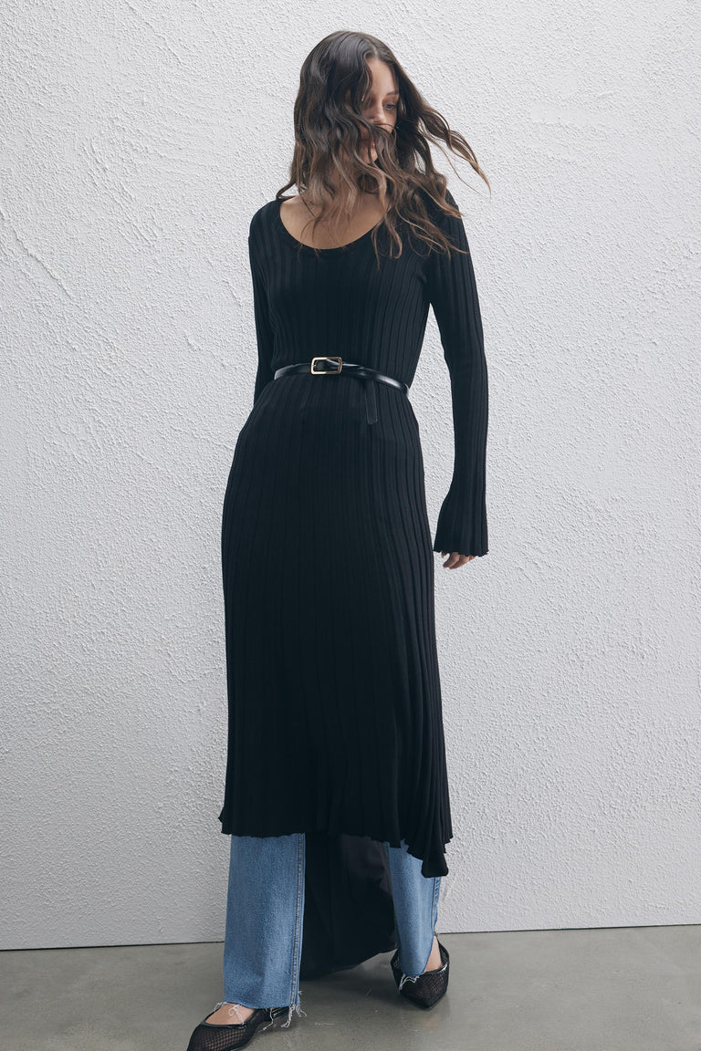 Woman wearing a black ribbed knit scoop neck dress with long sleeves, styled with a belt and layered over blue jeans