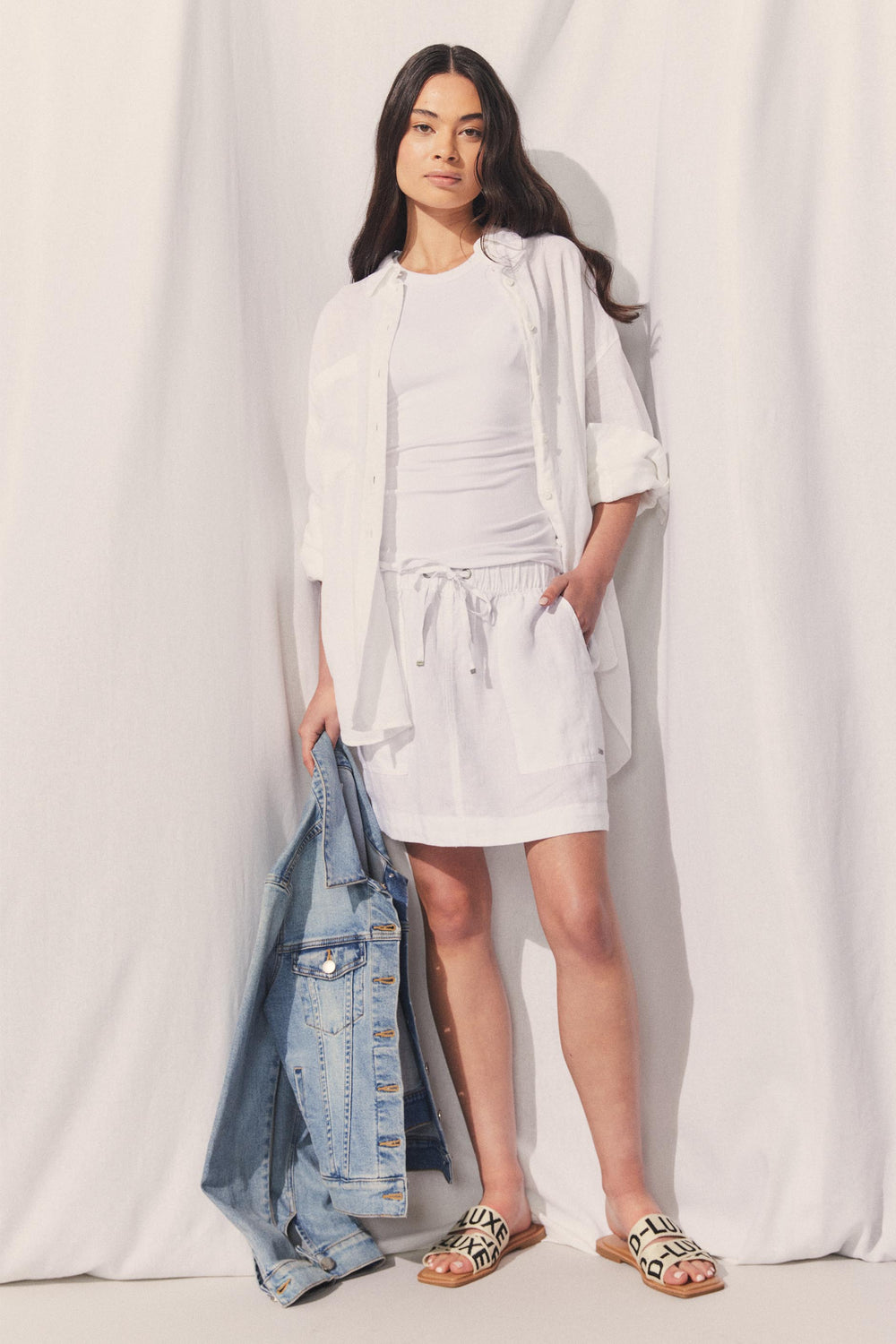 Model wearing D-Luxe Linen Mini Skirt with elastic waistband and side pockets, holding denim jacket in portrait view.